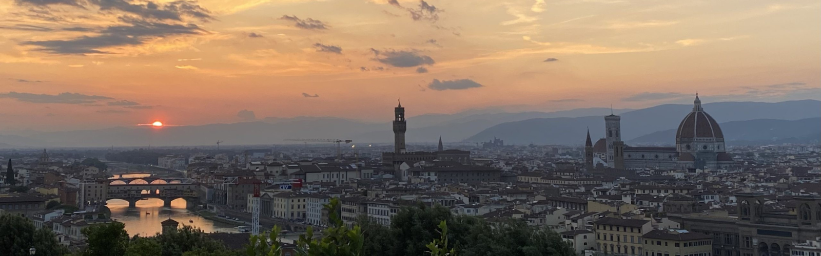 The many tan, brick buildings with reddish-brown roofs of the city of Florence, including the tall, domed Duomo, are illuminated by the setting sun. 